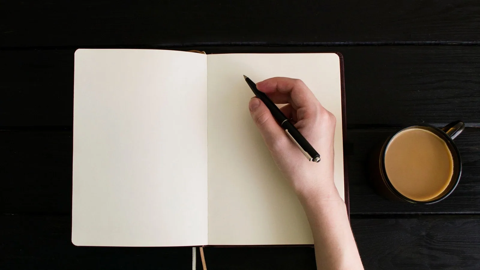 A hand writing with a black pen in an open blank notebook beside a cup of coffee on a dark table
