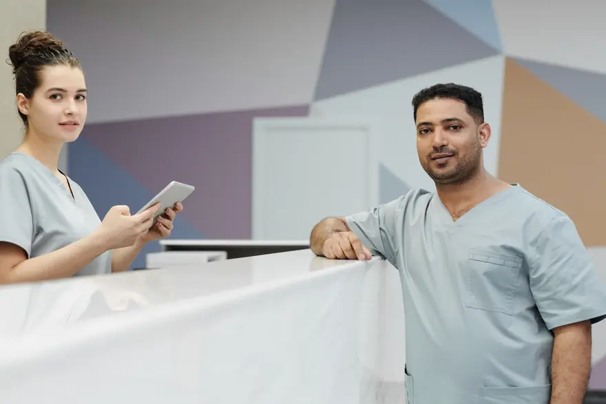 Two clinic staff in scrubs at a reception desk, one holding a tablet