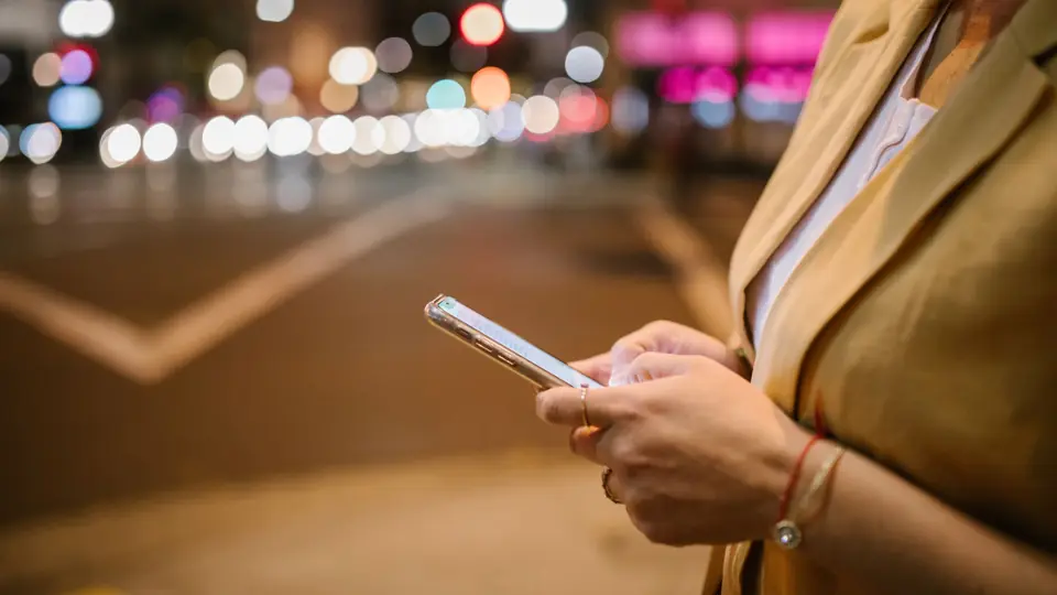 Someone checks a smartphone on a city street at night, with lights blurred behind