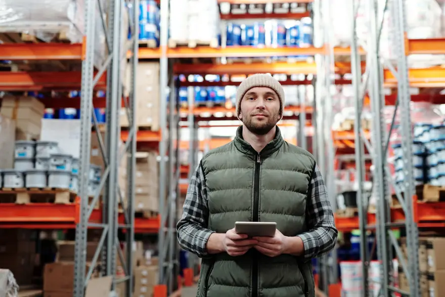 A warehouse worker holding a tablet between tall shelves of stock
