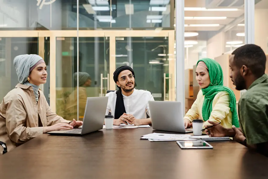 A small team in headscarves and casual clothes works on laptops around a meeting table