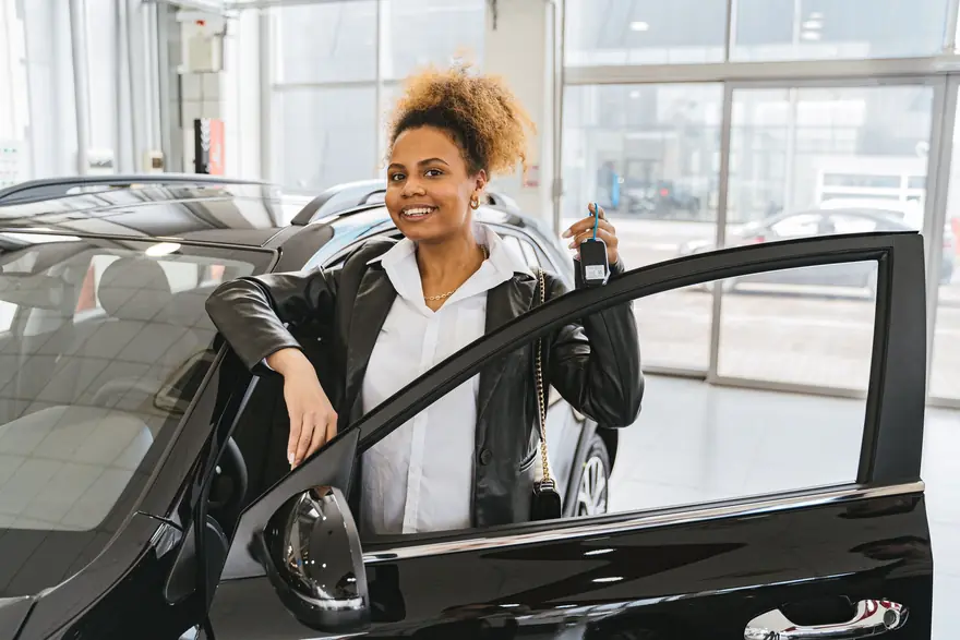 A smiling woman holds up a car key beside the open door of a new car in a showroom