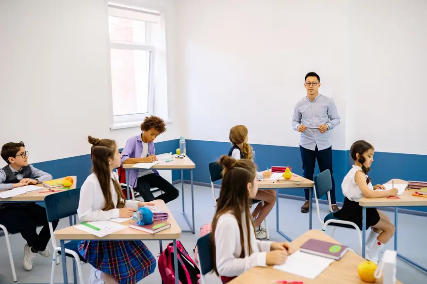A teacher stands at the front of a bright classroom as pupils work at their desks
