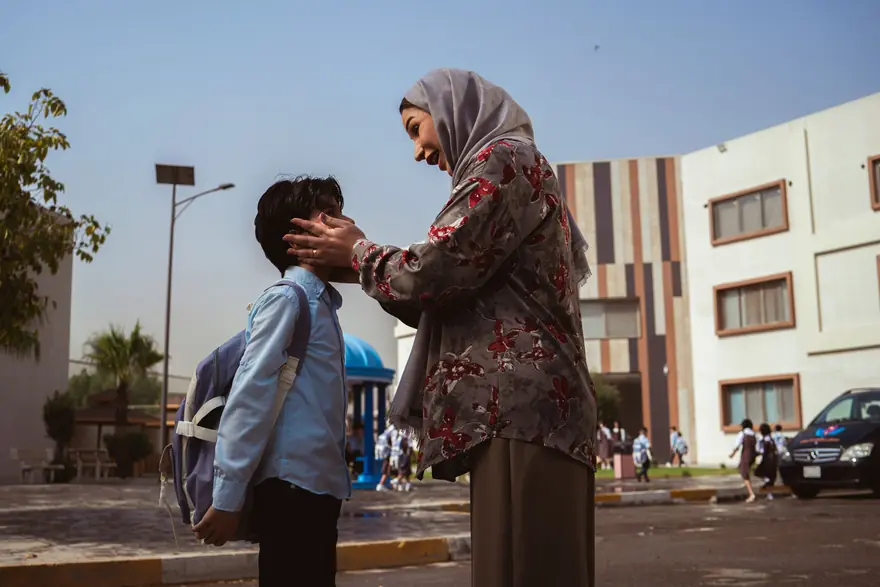 A mother in a headscarf says goodbye to her son with a school bag outside a school