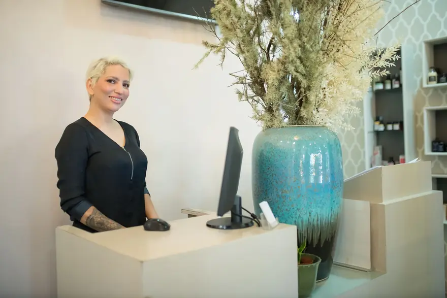 A smiling receptionist stands behind a clinic desk beside a large blue vase
