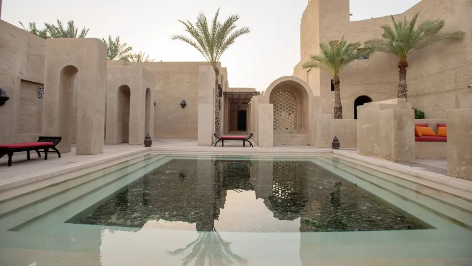 A still pool in the stone courtyard of a desert resort, framed by palm trees and arches