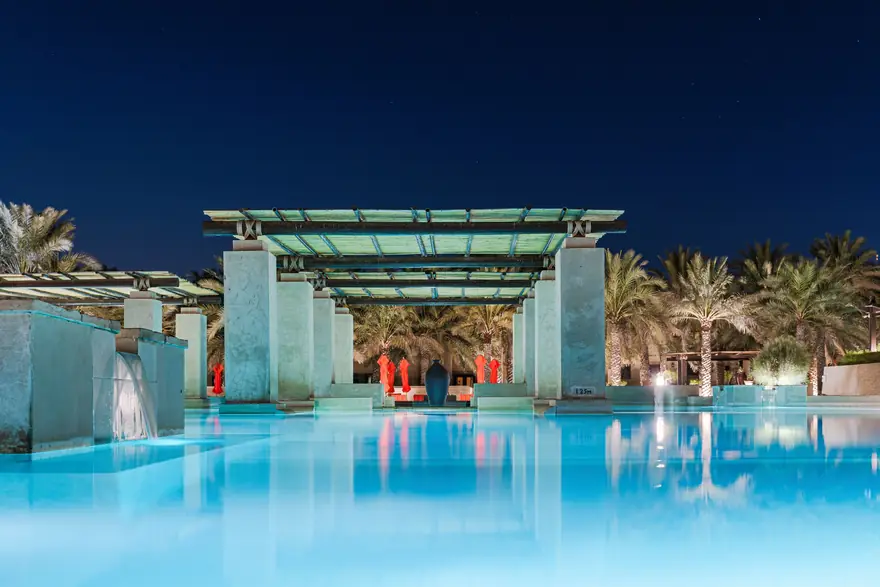 A resort swimming pool at night under a lit pergola, with palm trees behind