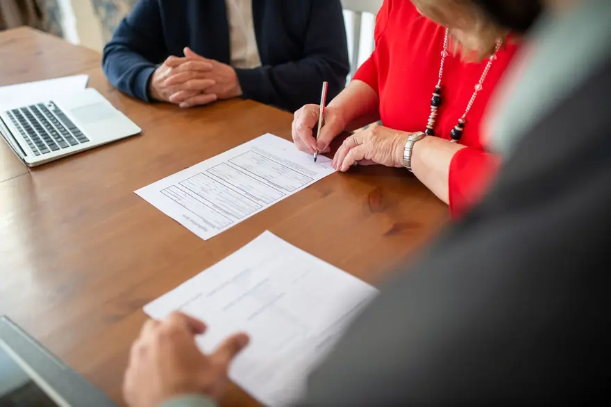 A woman signs a form at a wooden table while an adviser looks on