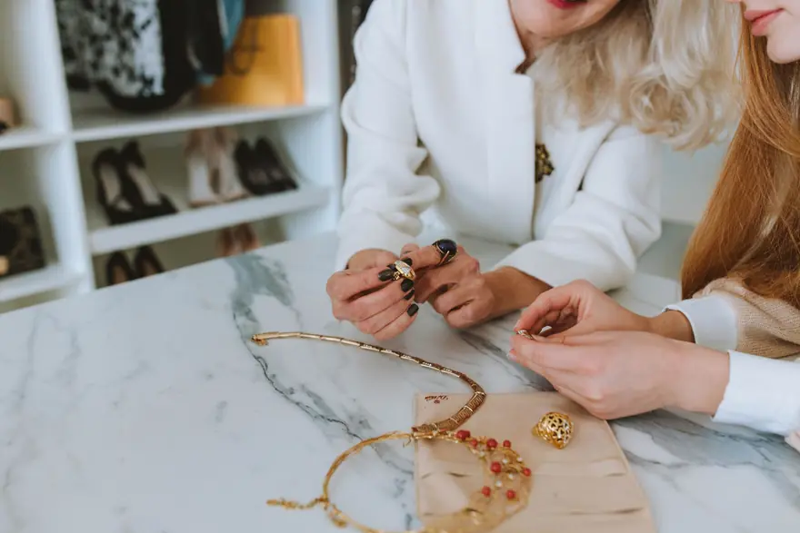 Two women look closely at gold jewellery laid out on a marble table