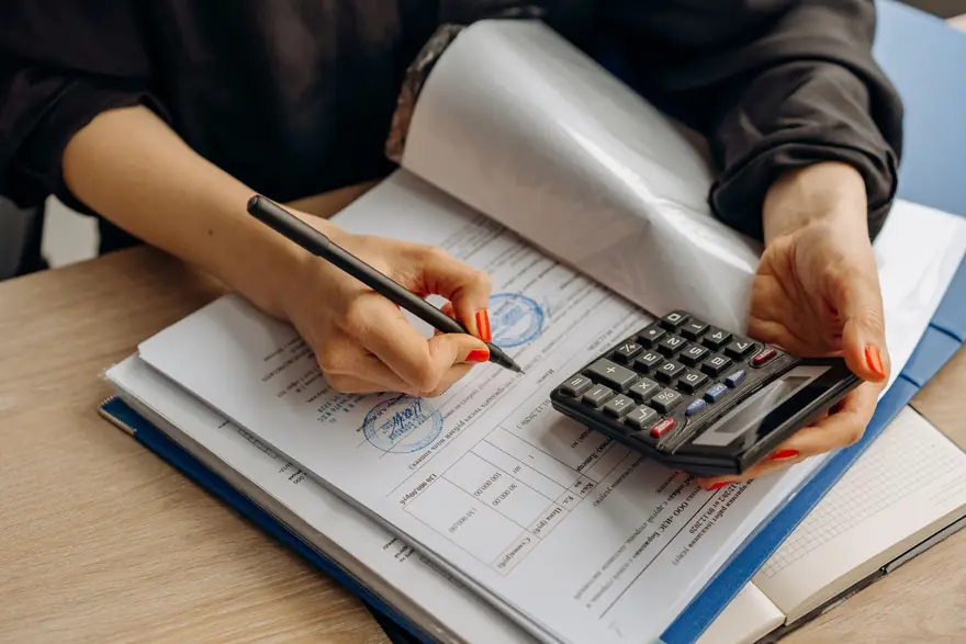 A hand holds a pen over stamped forms beside a calculator