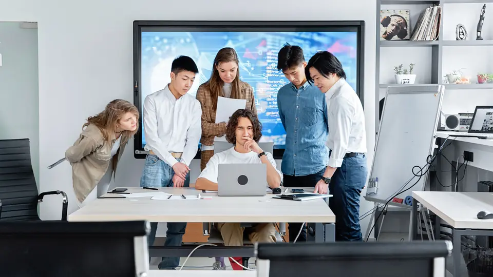 A small team gathers around a laptop in a bright office in front of a large screen