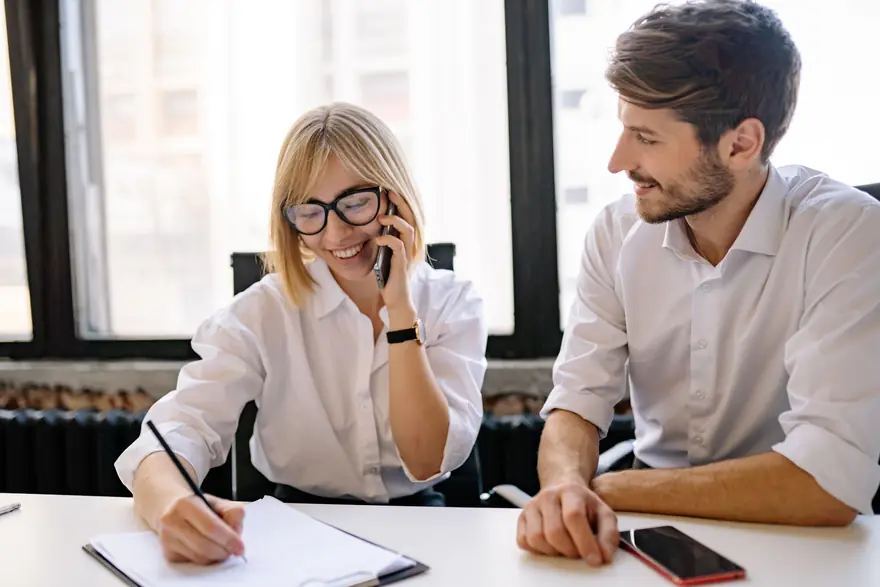 A woman takes a phone call and writes notes while a colleague beside her looks on