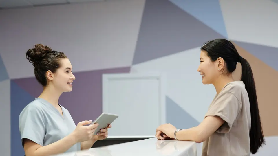 A clinic receptionist holding a tablet talks with a smiling patient across the front desk