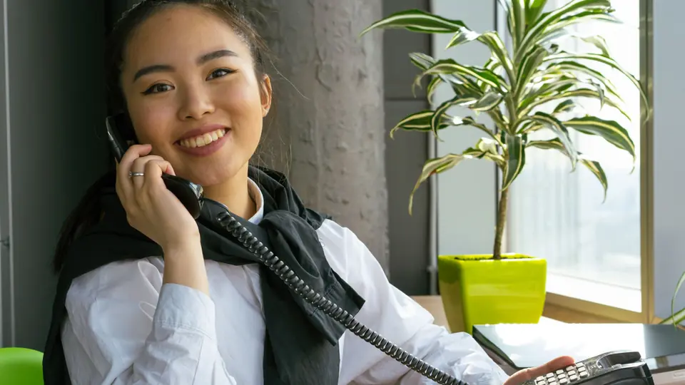 A smiling receptionist takes a call on a desk phone beside a window