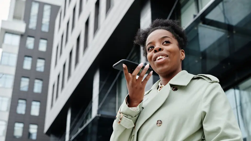A woman in a green coat records a voice message on her phone outside an office building