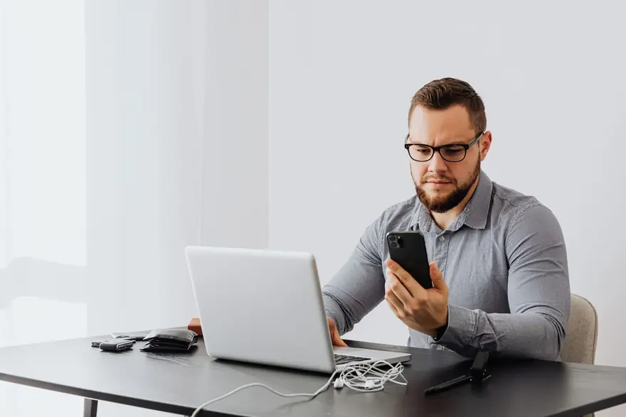 A man at a desk with a laptop reads a message on his phone
