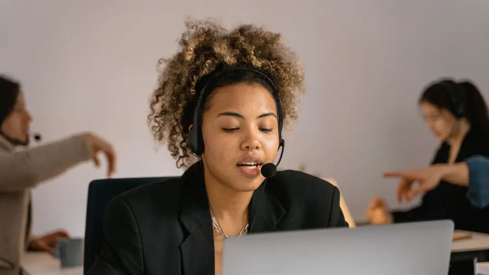 A woman wearing a headset speaks while looking at a laptop in an office