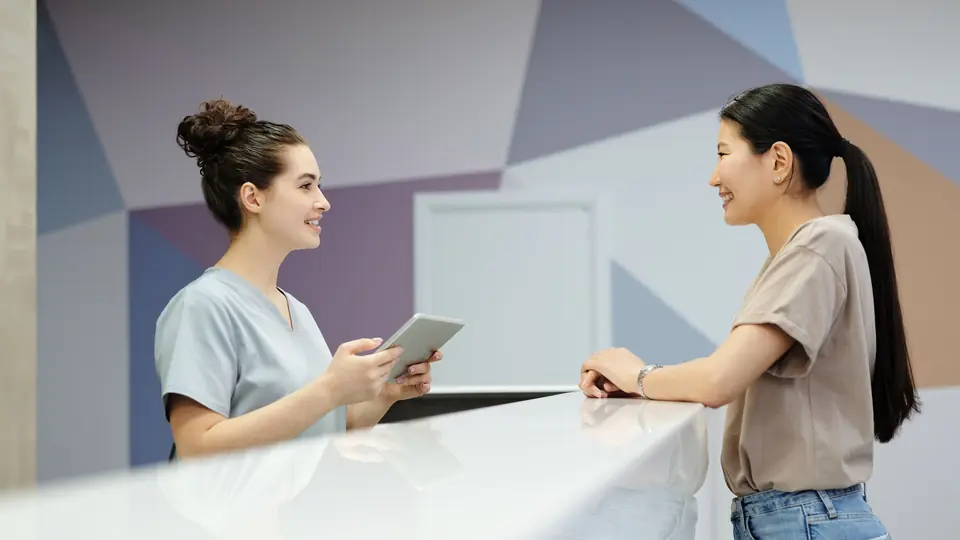 A receptionist holding a tablet talks with a patient across a white clinic desk