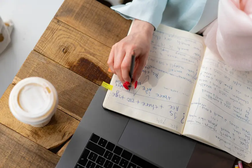 A hand writes notes in an open notebook next to a laptop and a coffee
