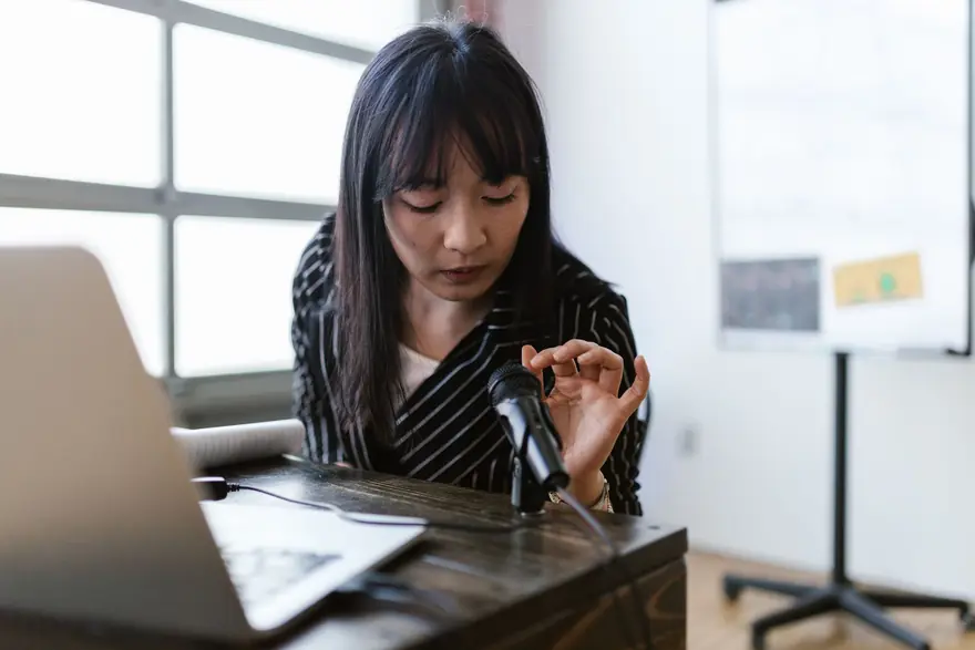 A woman adjusts a small microphone on a desk beside a laptop