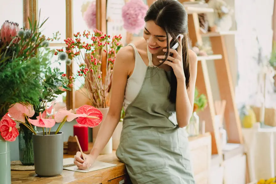 A florist takes a call at the counter of a flower shop while writing a note