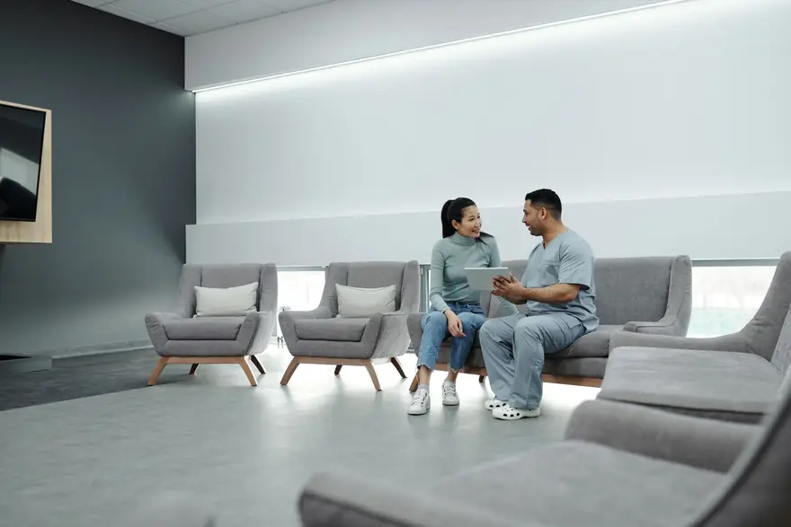 A nurse and a patient talk on a sofa in a bright clinic waiting area