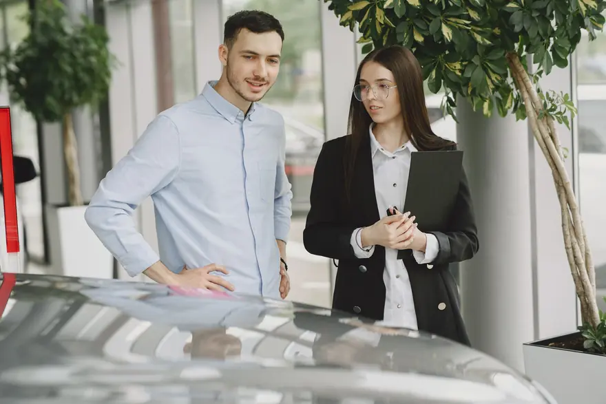 A salesperson with a folder talks with a customer beside a car in a showroom