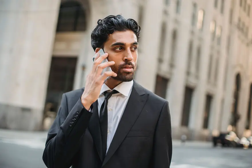 A young man in a dark suit takes a phone call on a city street