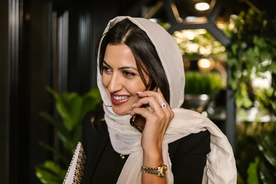 A smiling woman in a cream headscarf talks on her phone in a softly lit lounge