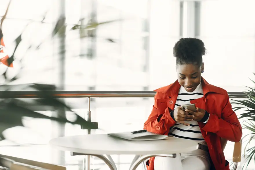 A woman in a red jacket reads her phone at a table in a bright cafe