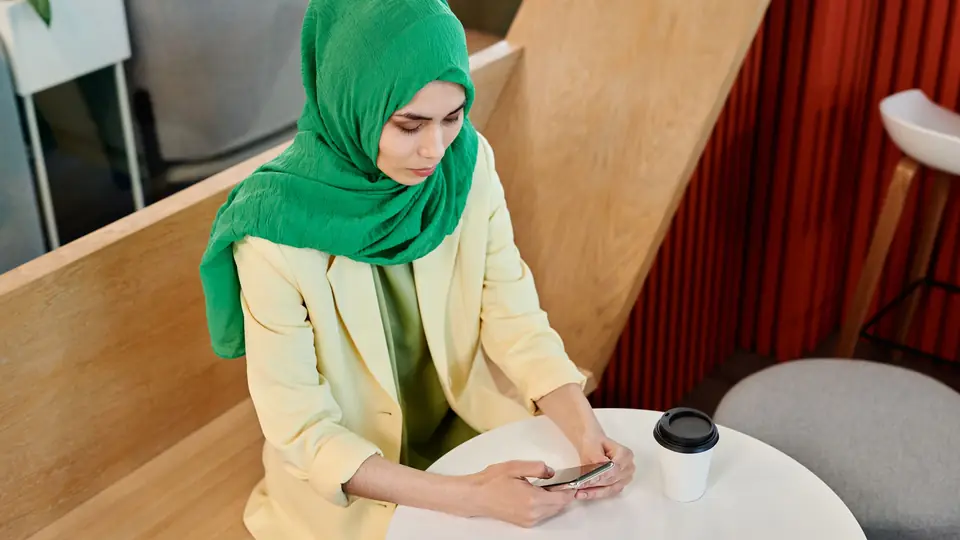 A woman in a green headscarf reads her phone at a cafe table