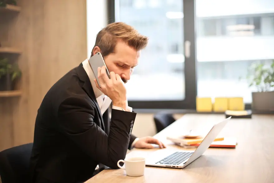 A man in a suit takes a phone call at his desk beside a laptop