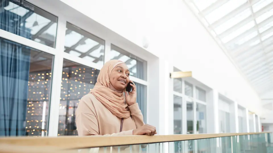 A woman in a beige headscarf smiles as she talks on her phone in a bright glass atrium