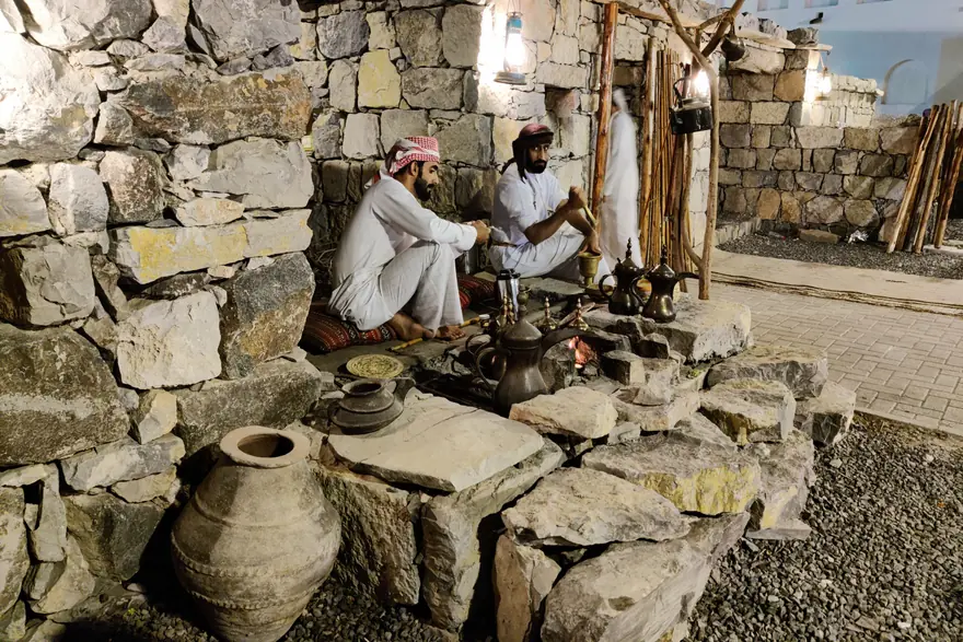 Two men in traditional dress sit beside coffee pots at a stone heritage courtyard