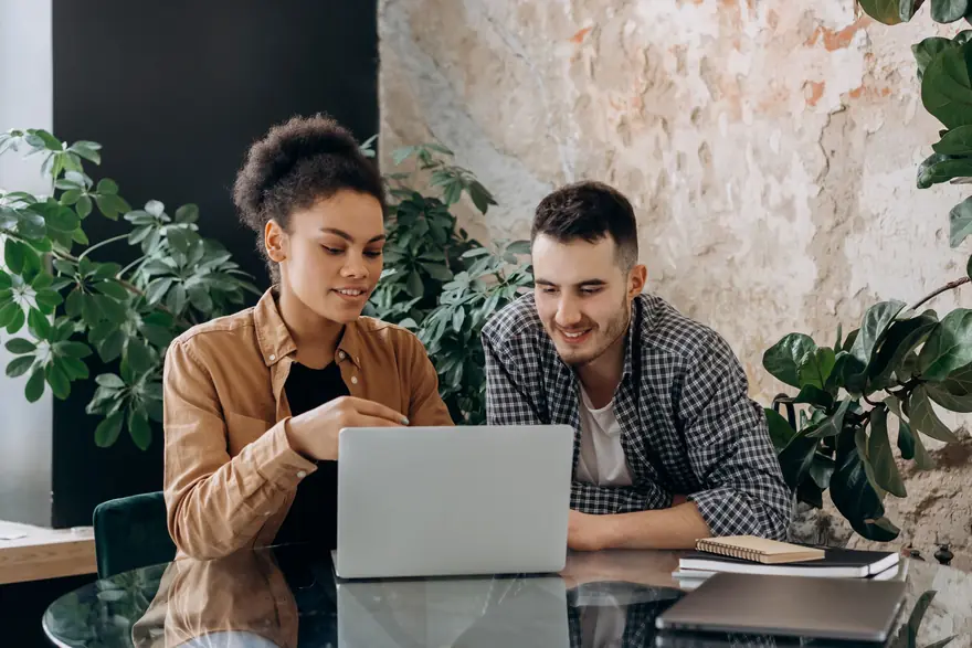 Two colleagues look at a laptop together in a bright office with plants