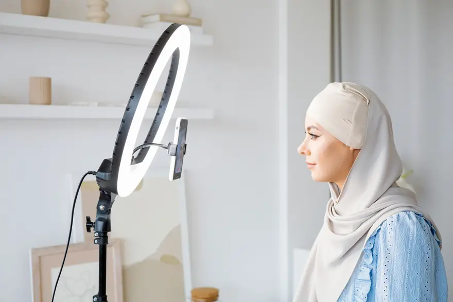 A woman in a white headscarf sits beside a ring light set up for filming