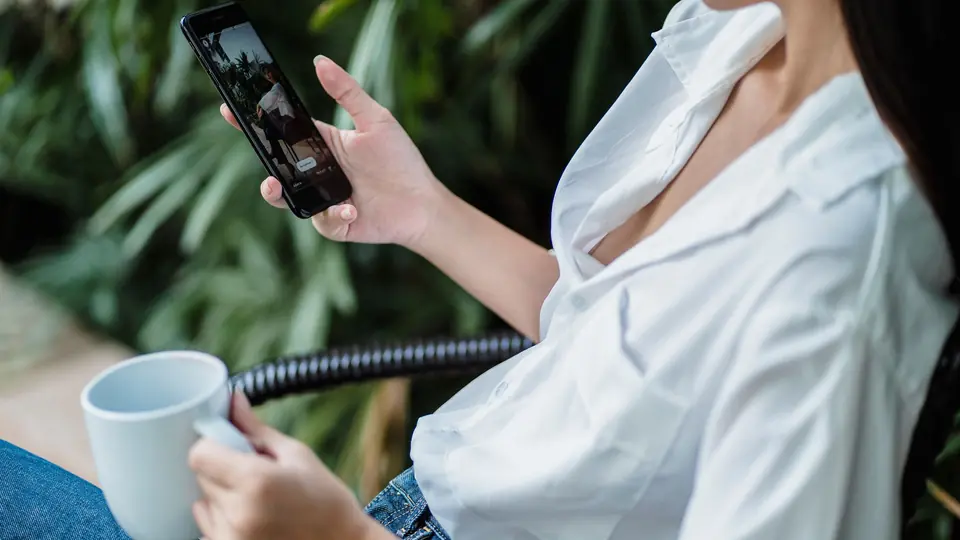A woman in a white shirt scrolls on her phone holding a mug, plants behind her