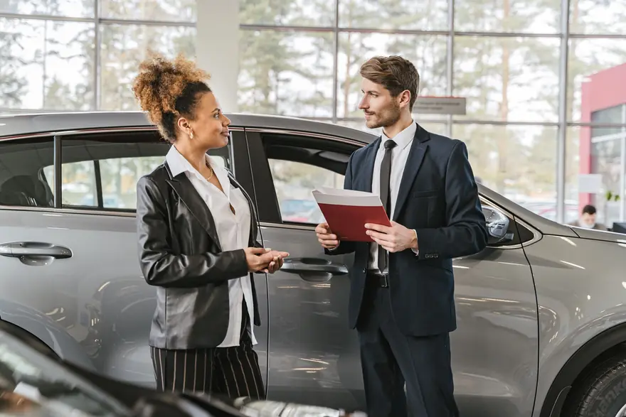 A salesperson holding a folder talks with a customer beside a car in a showroom