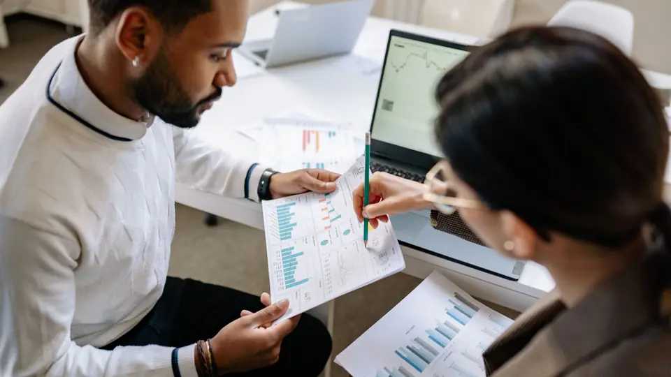 Two colleagues review printed charts beside a laptop showing a graph
